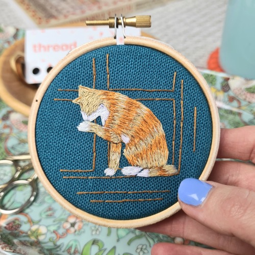 Needle painting embroidery of ginger-and-white tabby cat licking its paw on blue fabric in an embroidery hoop