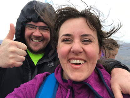wind-swept brunette woman and man in hooded raincoat giving a thumbs up