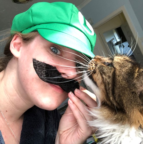 Woman wearing green cap and fake black moustache with tabby-and-white cat