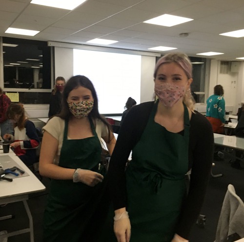 one long-haired brunette woman and one long-haired blonde woman both wearing face masks and green aprons inside university lecture room