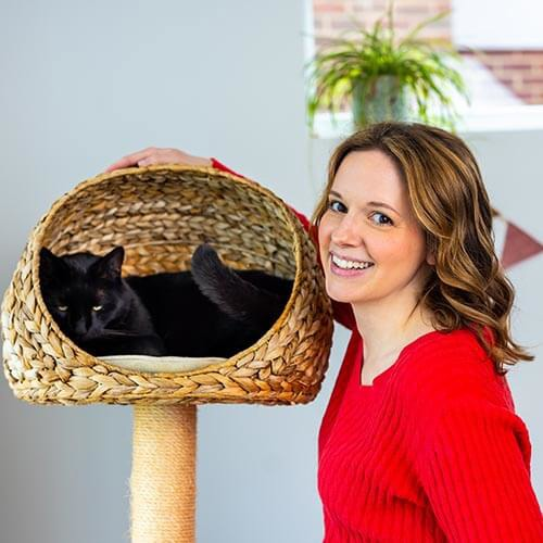 Women with long brunette hair wearing red top standing next to a wicker cat bed on top of a cat tower. A black cat is sleeping in the cat bed