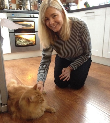 Woman with long blonde hair and wearing a blue-and-grey stripy top kneeling on the  the wooden kitchen floor stroking a long-haired ginger tabby cat
