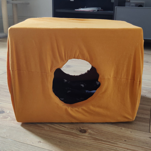 A cardboard box covered with a yellow t-shirt to make a cat tent. There is a hole in the front and back of the box and a blanket inside