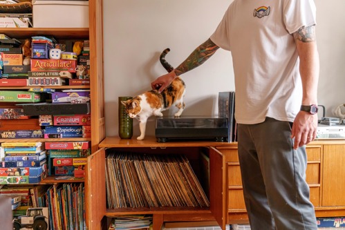 A tortoiseshell-and-white cat walking across a covered vinyl record player and being stroked by a man with tattoos on his arm and wearing a white t-shirt and grey trousers. A shelf of vinyl records is below the record player